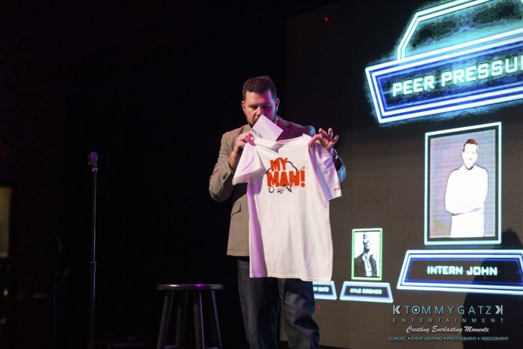 A man stands on stage holding a white T-shirt with the words "MY MAN!" printed on it. A microphone stand and a screen displaying "PEER PRESSURE" are in the background.