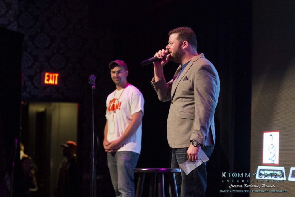 Two people on stage; one speaking into a microphone, the other standing near a stool. Exit sign visible in the background. Audience partially visible.