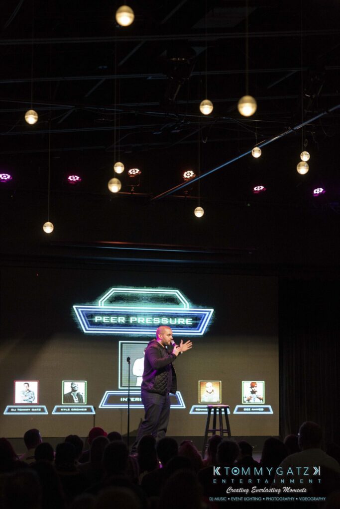 A person on stage presents a game show titled "Peer Pressure" with a futuristic backdrop and illuminated hanging lights above.