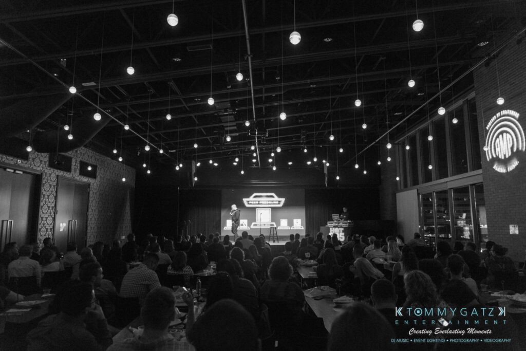 A speaker addresses an audience in a dimly lit venue with hanging lights and a large screen displaying a logo on stage.