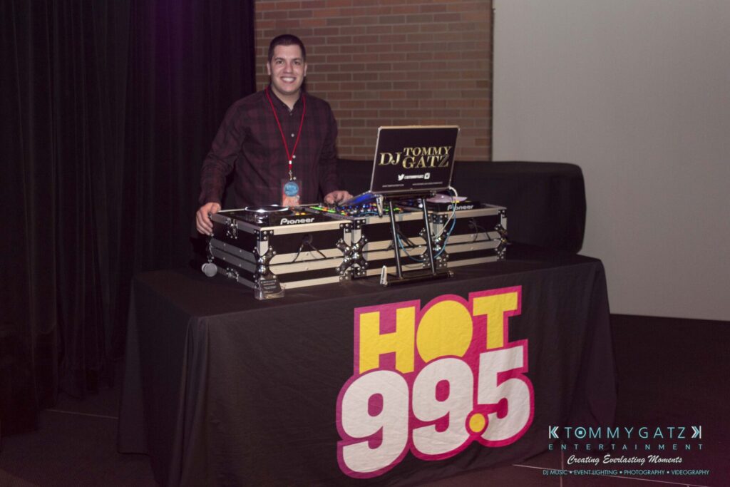A DJ stands behind a booth with equipment, featuring a "Hot 99.5" banner.