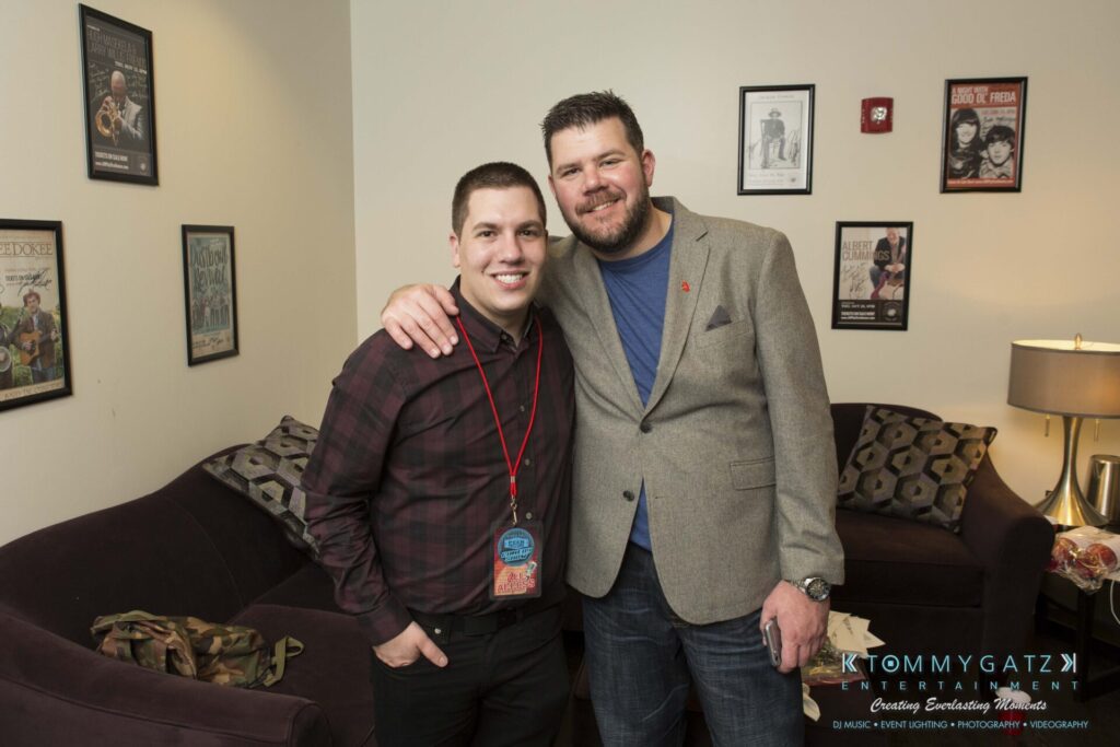 Two men posing together in a living room with framed posters on the wall behind them. One wears a plaid shirt and lanyard, the other a gray blazer. A couch and a standing lamp are visible.