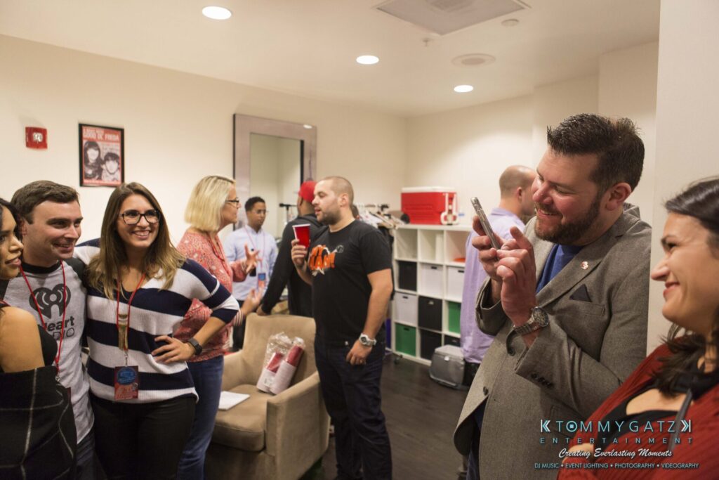 A group of people socializing indoors, with some holding drinks and one person taking a photo with a smartphone.