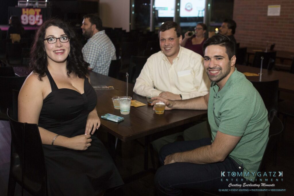 Three people sitting at a wooden table with drinks, smiling at the camera in a dimly lit venue.