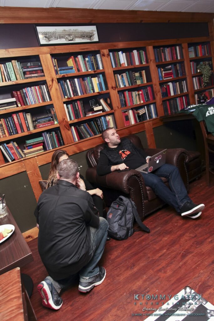 Three people in a room with bookshelves. Two seated on a couch, one on the floor, with a laptop. Backpack on the floor, and a table with food nearby.