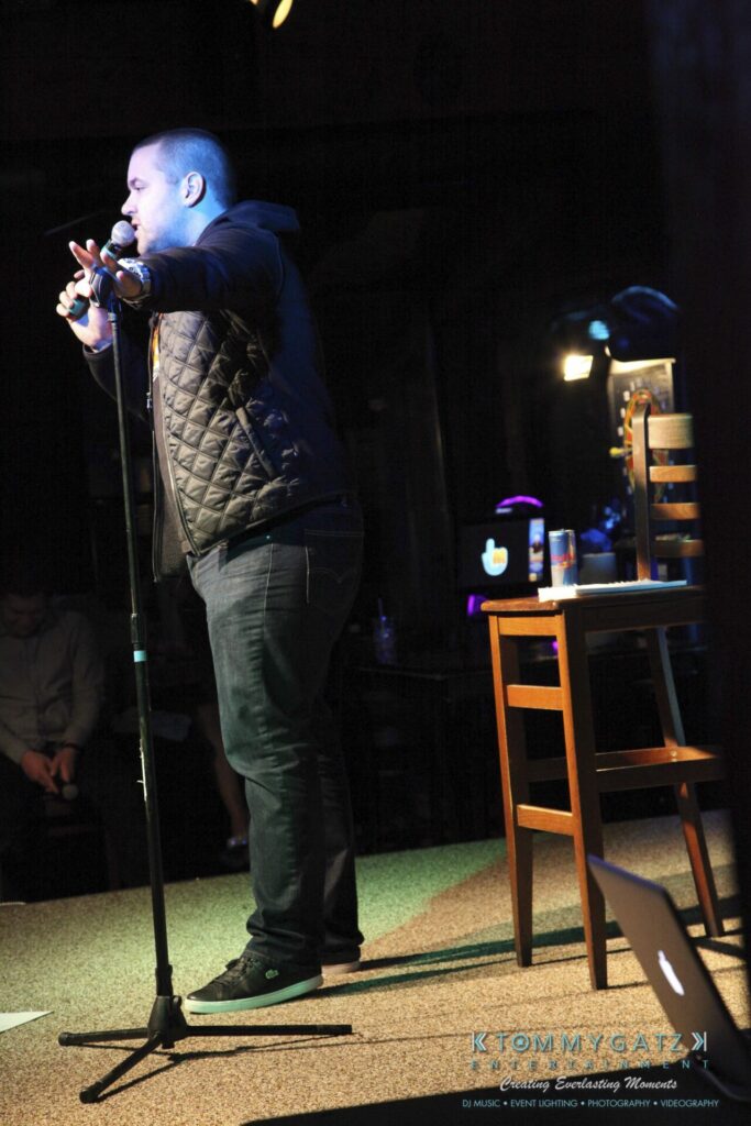 Comedian performing on stage, holding a microphone. A chair and table with a drink are in the background.