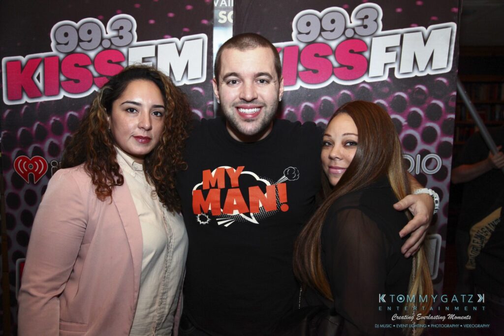 Three people posing in front of a "99.3 KISS FM" backdrop. The man in the center is wearing a black "My Man!" t-shirt.