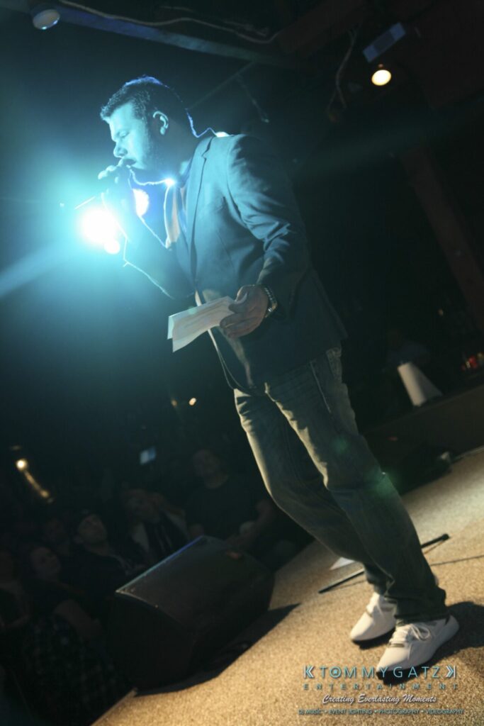 A man in jeans and a jacket speaks into a microphone on stage in front of an audience, illuminated by bright stage lights.
