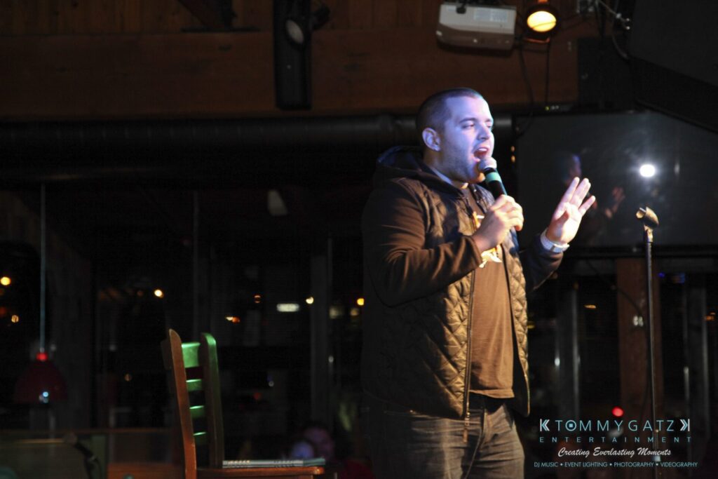 A person is speaking into a microphone on a dimly lit stage with a chair nearby.