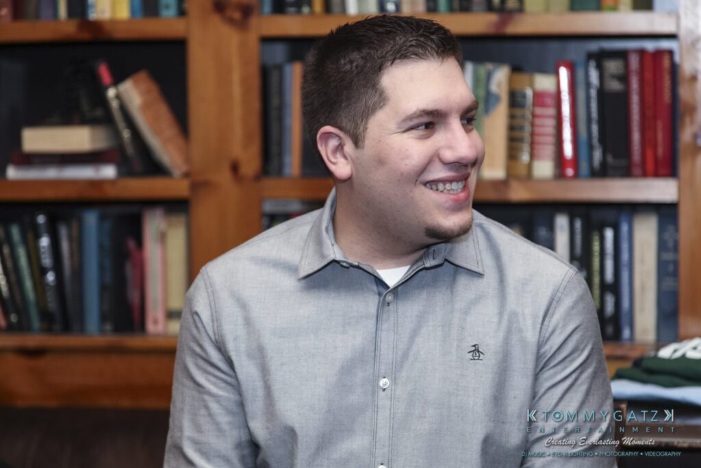 A person wearing a gray button-up shirt smiles while seated in front of a wooden bookshelf filled with books.