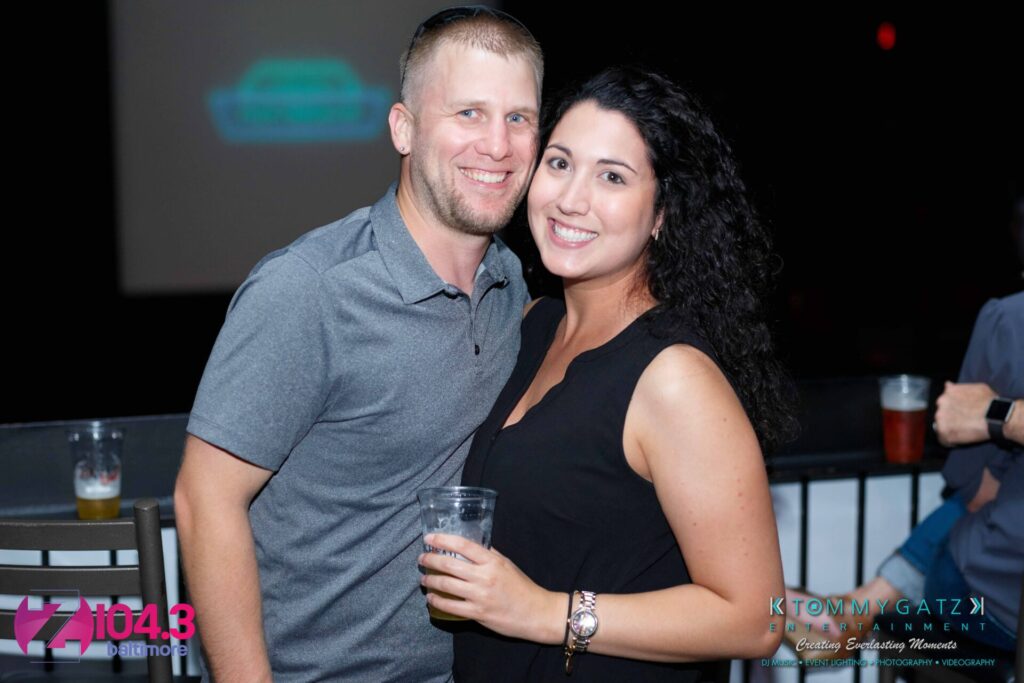 A smiling man and woman stand close together, holding drinks, at an event. Logos for "Z 104.3 Baltimore" and "Tommy Gatz Entertainment" are in the lower corners.