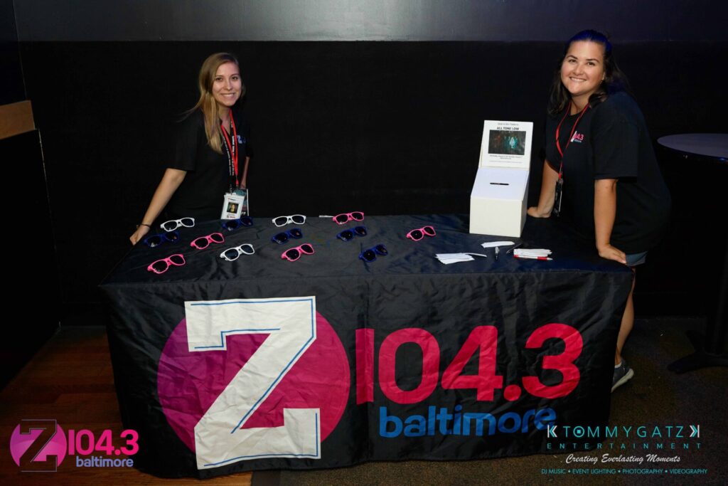 Two people stand behind a table with Z104.3 Baltimore merchandise, including sunglasses and lanyards, at an event.