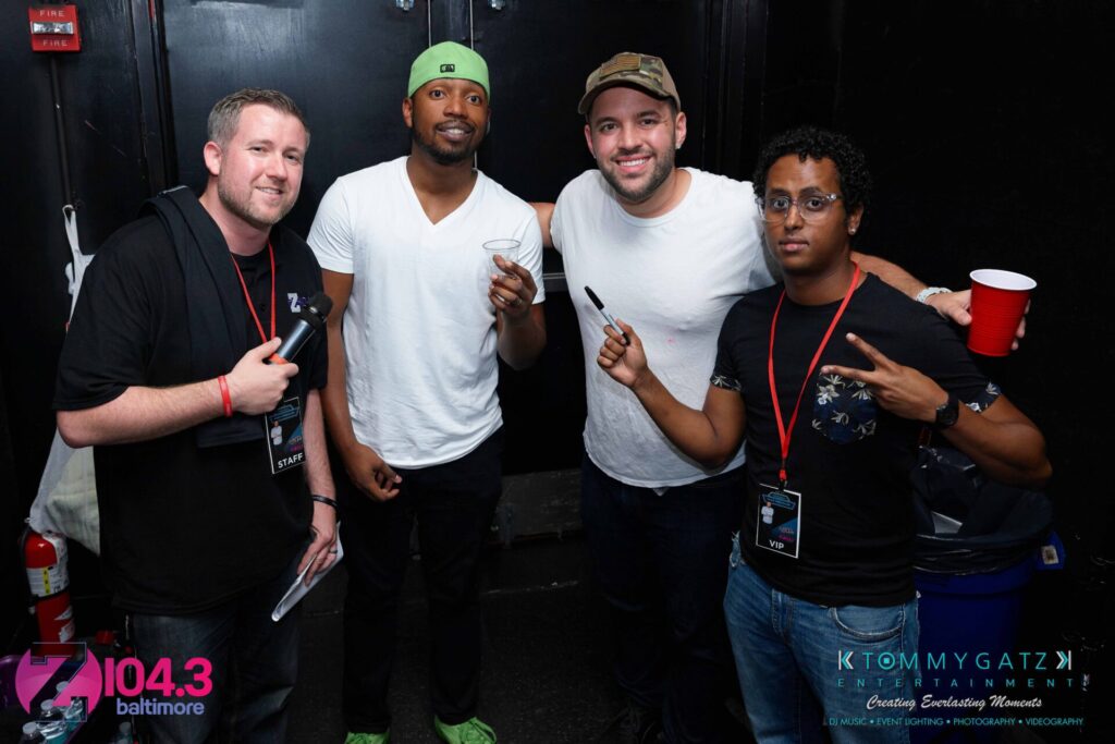 Four men posing together, two holding drinks. They are in a room with black walls, and one wears a green cap. A logo and text about Baltimore's 104.3 radio station is visible in the corner.