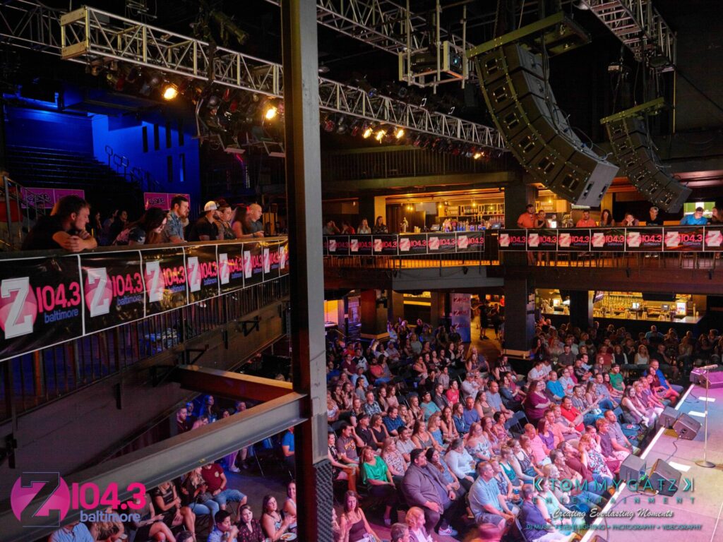 An indoor concert venue with a crowded audience seated in front of a stage. Banners for "Z104.3 Baltimore" are displayed throughout the venue.