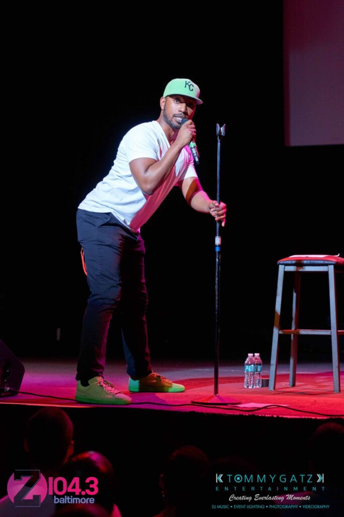 A performer in a white shirt and green cap sings into a microphone on a stage with a stool nearby, under colorful lighting. Logos of "Z104.3 Baltimore" and "Tommy Gatz Entertainment" are visible.