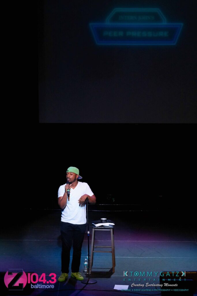 A man wearing a cap is speaking on stage in front of a stool and mic stand, with a Z104.3 Baltimore logo in the bottom corner.