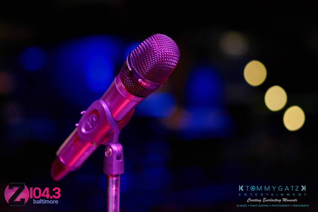 A microphone on a stand with blurred lights in the background. Logos for Z104.3 Baltimore and Tommy Gatz Entertainment are visible.