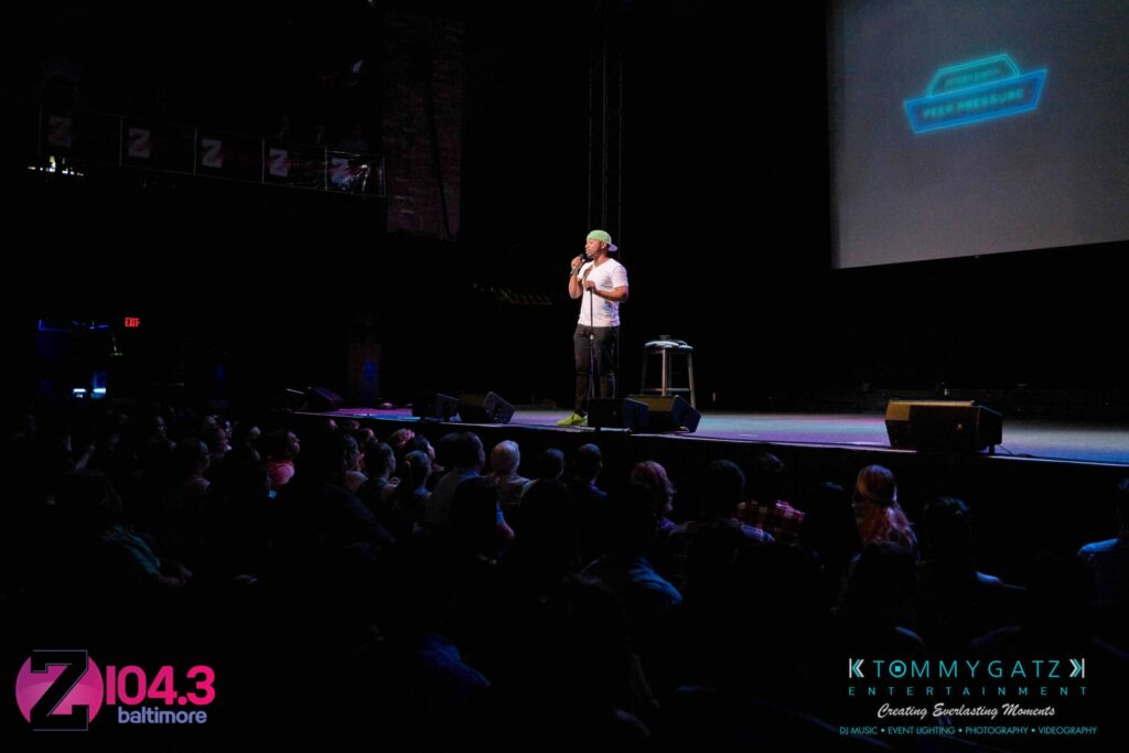 A person stands on stage performing in front of a seated audience, with logos and company names visible on stage and the foreground.