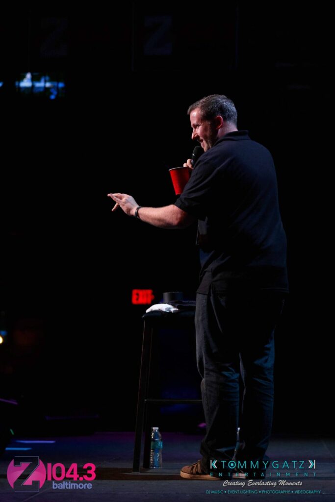 A man stands on stage holding a red cup and microphone, facing an audience. There's a stool and bottle of water nearby.