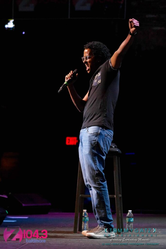 A man stands on stage holding a microphone, gesturing passionately. He is next to a stool with two water bottles on the floor. Banners for Z104.3 Baltimore are in the background.