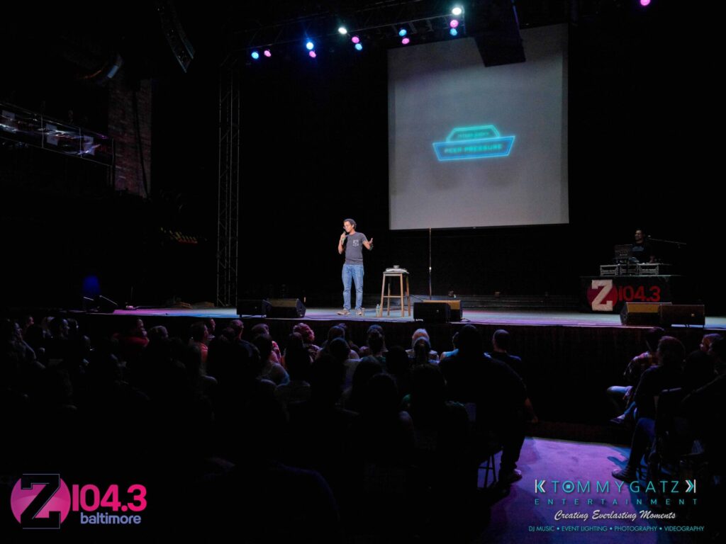 Comedian performing on stage with a microphone, audience seated in front. Bright stage lights and a screen displaying text behind the performer. Radio station logo visible on stage.