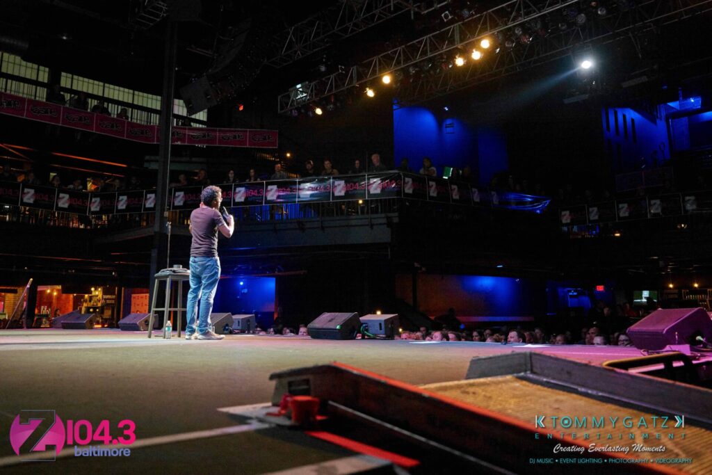 Comedian performing on stage at a venue with a seated audience. Stage lights are focused on the performer, and a large banner in the background displays the radio station Z104.3 Baltimore.