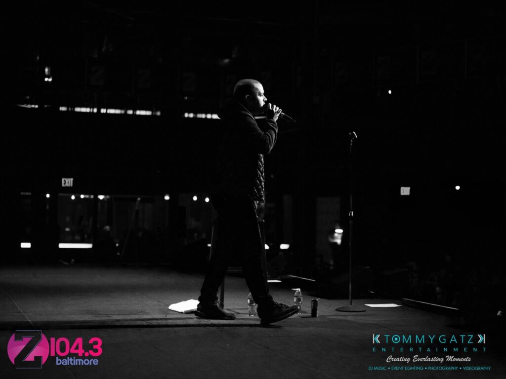 A person on stage holds a microphone, performing in front of a dark audience. Logos for Z104.3 Baltimore and Tommy Gatz Entertainment are visible.