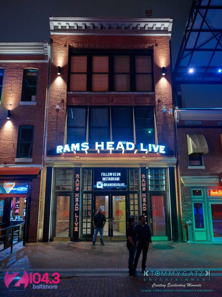 Exterior of Rams Head Live venue at night, with neon signage and a digital display above the entrance. Two people stand outside near the entrance.
