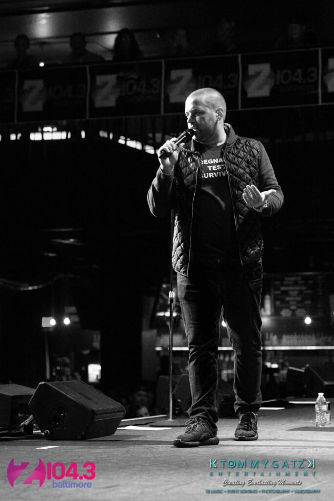 A person stands on stage holding a microphone, speaking to an audience. The background features branding for a radio station.