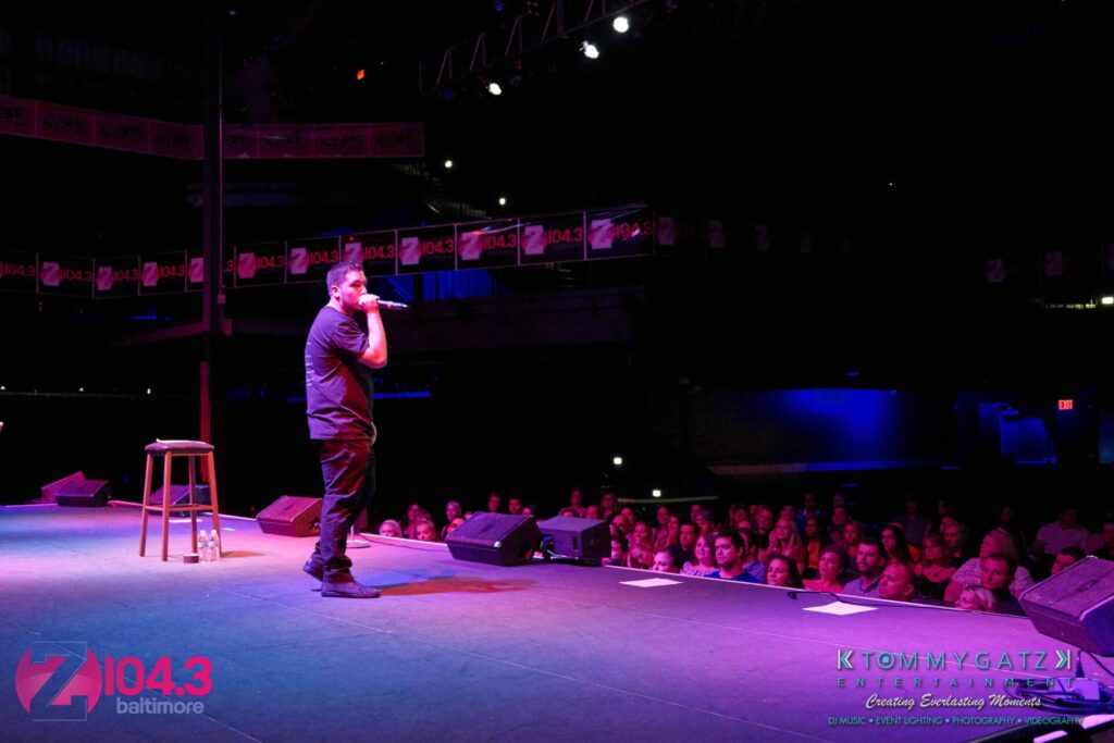 Comedian performing on stage in front of a seated audience, with a stool beside him. Stage banners and lighting visible.