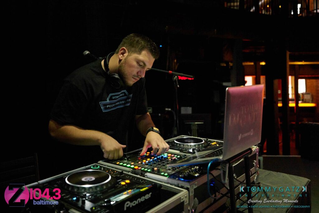 A DJ with headphones operates music equipment on a dimly lit stage. Multiple logos are visible in the scene.