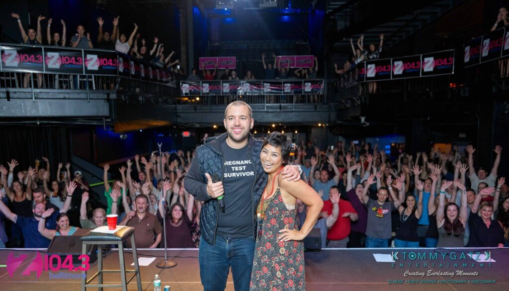 Two people pose on stage with a cheering crowd in the background. A banner displays "Z104.3 Baltimore.