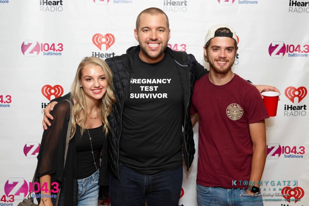 Three people pose in front of a backdrop with logos, including iHeart Radio and Z104.3. The center person wears a shirt reading "Pregnancy Test Survivor.