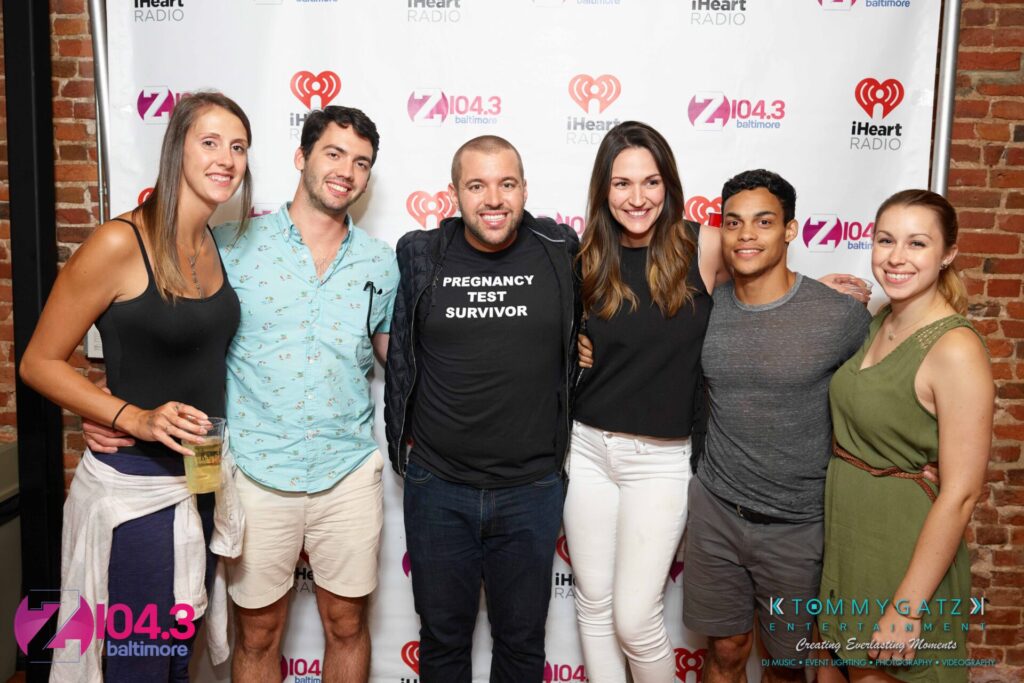 A group of six people posing together in front of a backdrop. One person in the center wears a "Pregnancy Test Survivor" shirt. Logos for Z104.3 and iHeart Radio are visible.