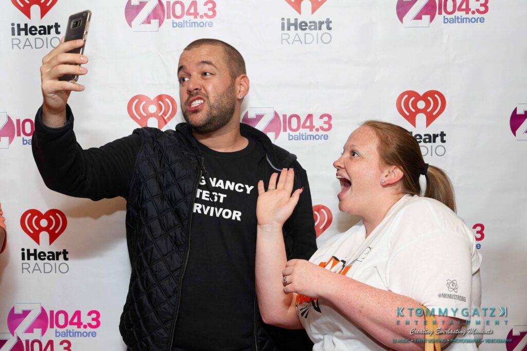 Two people taking a selfie in front of a backdrop with radio station logos.