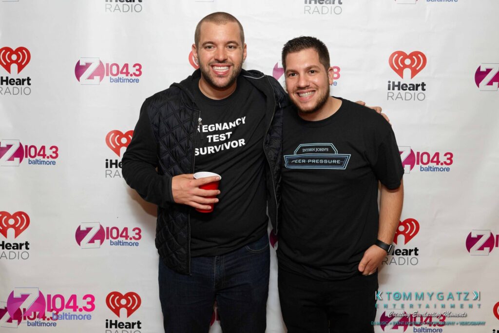 Two men posing together at an event, standing in front of a backdrop featuring logos of Z104.3 Baltimore and iHeartRadio. One is holding a red cup.