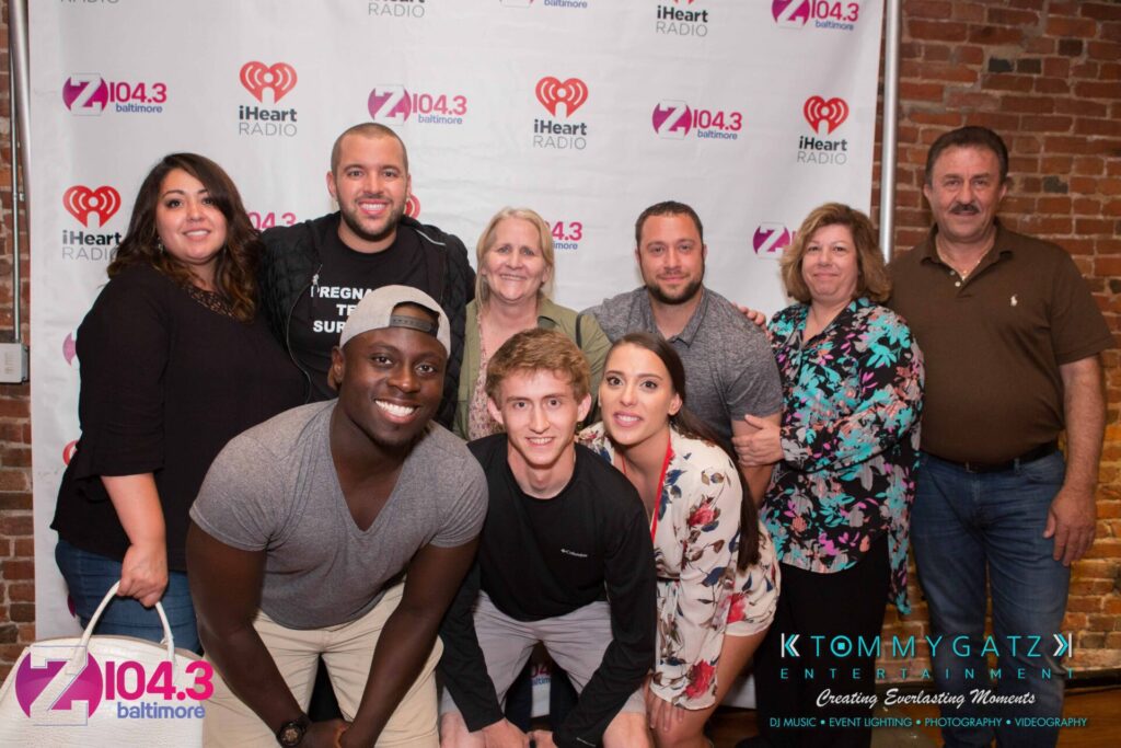 A group of people posing together in front of a step-and-repeat banner with various logos, including a radio station and entertainment company.