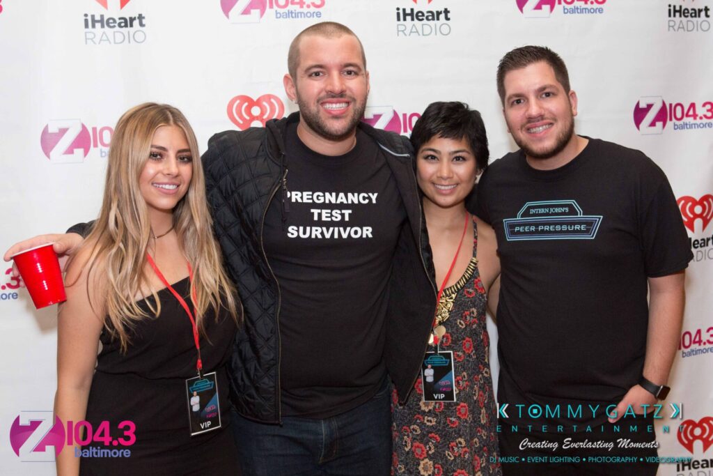 Four people posing in front of a step-and-repeat banner with logos from iHeart Radio and Z104.3 Baltimore. One person holds a red cup. Two are wearing VIP lanyards.