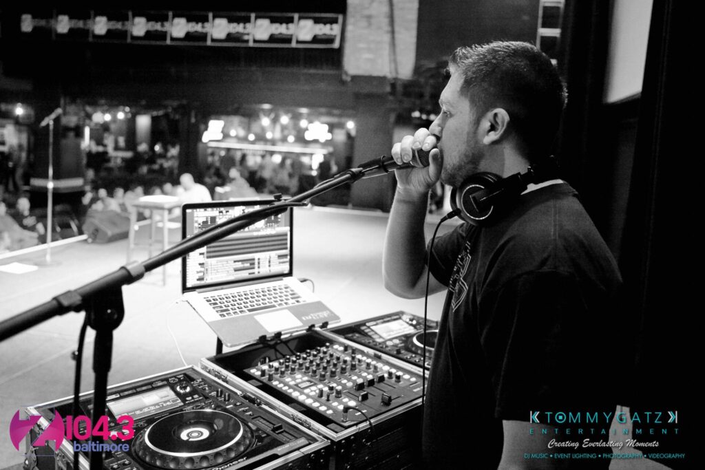 A DJ is performing on stage, holding a microphone, with music equipment in front of him and a laptop open to his right.