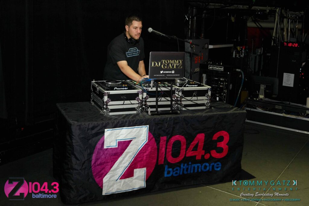A DJ stands behind a table with audio equipment, branded with "Z104.3 Baltimore" and "DJ Tommy Gatz." The setting appears to be a dimly lit indoor venue.