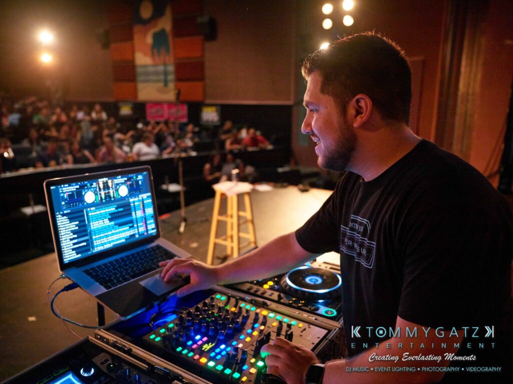A DJ is performing on stage with a laptop and mixer, surrounded by audience members in a dimly lit venue.