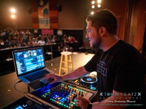 A DJ is performing on stage with a laptop and mixer, surrounded by audience members in a dimly lit venue.