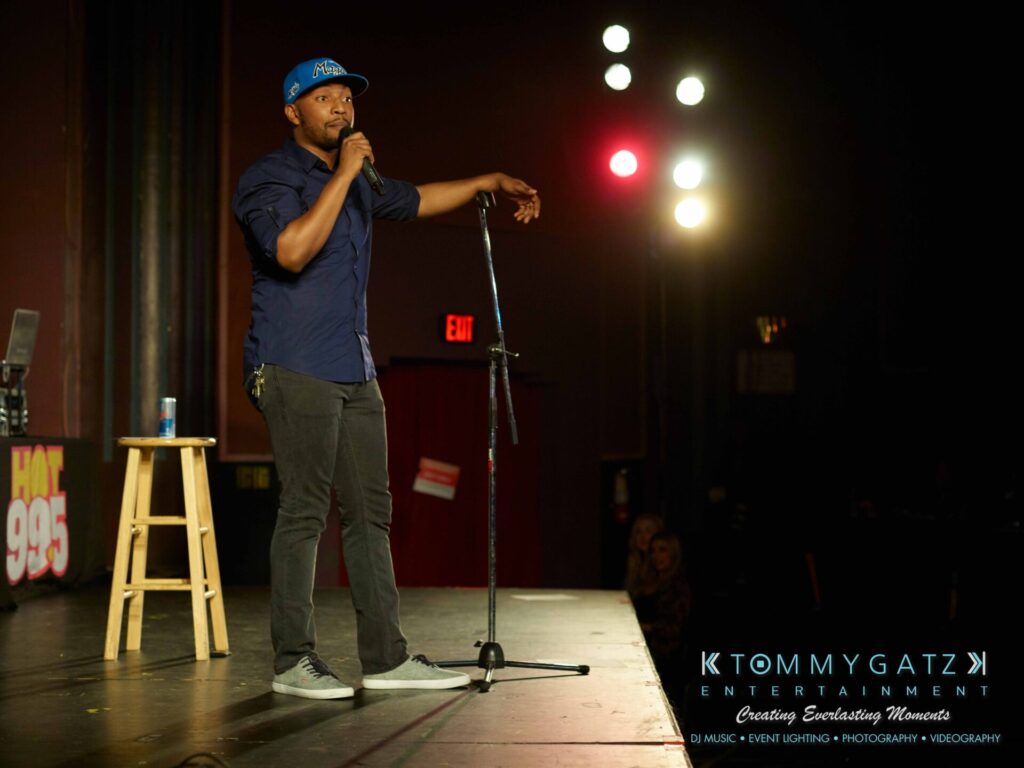 Comedian performing on stage with a microphone, stool, and drink can beside him. Bright lights illuminate the scene, and an audience is partially visible. Logo for Tommy Gatz Entertainment appears.
