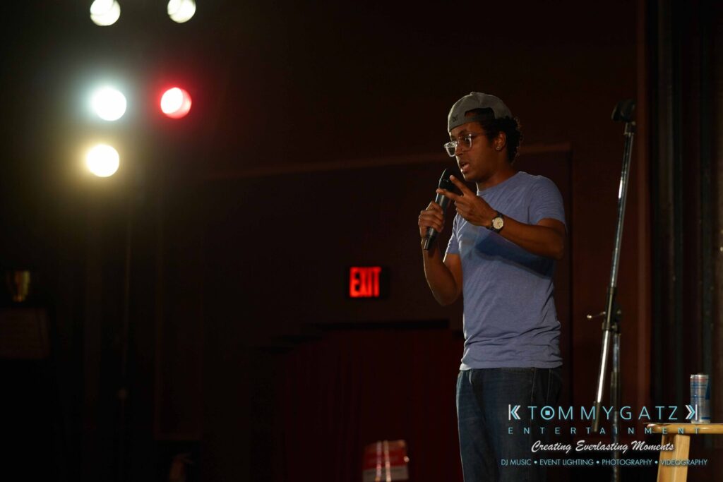 A person in a blue shirt and cap speaks into a microphone on stage, under bright lights, with "Tommy Gatz Entertainment" text overlay.