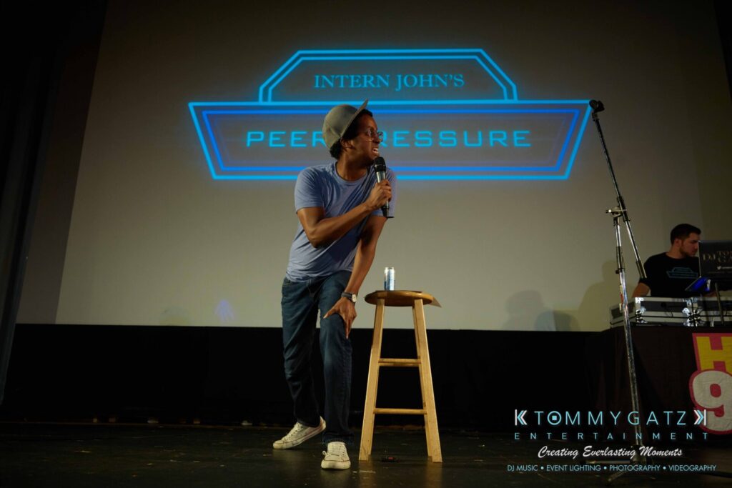 Person speaking into a microphone on stage next to a wooden stool, with a projected sign reading "Intern John's Peer Pressure" in the background.