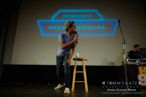 Person speaking into a microphone on stage next to a wooden stool, with a projected sign reading "Intern John's Peer Pressure" in the background.