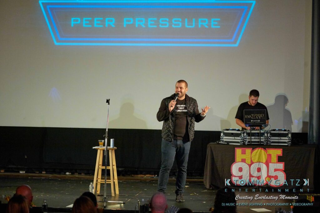 A man speaks into a microphone on stage with a DJ and a sign reading "Peer Pressure" above them.