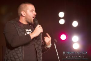 A man on stage holds a microphone, gesturing while speaking. Bright lights shine in the background.