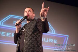 A man holding a microphone makes a gesture on stage, with illuminated text in the background.