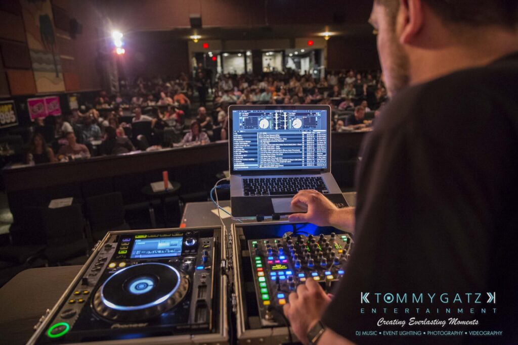 A DJ stands at a console with turntables and a laptop, mixing music for an engaged audience in a dimly lit venue.